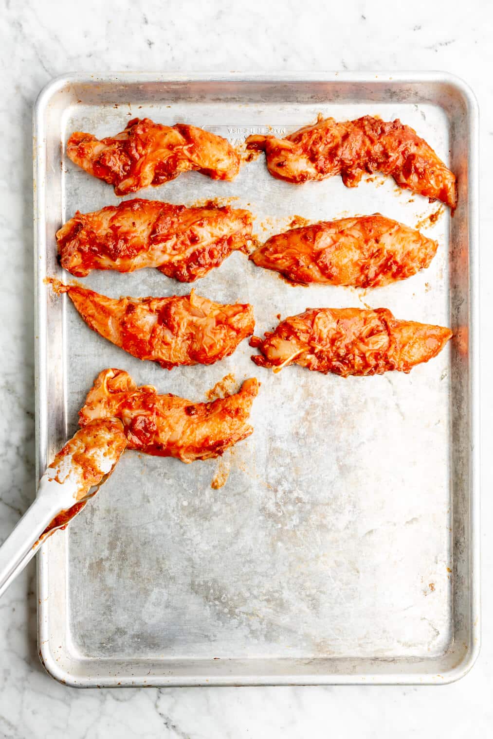 Tongs placing chicken coated in Tinga on a sheet pan
