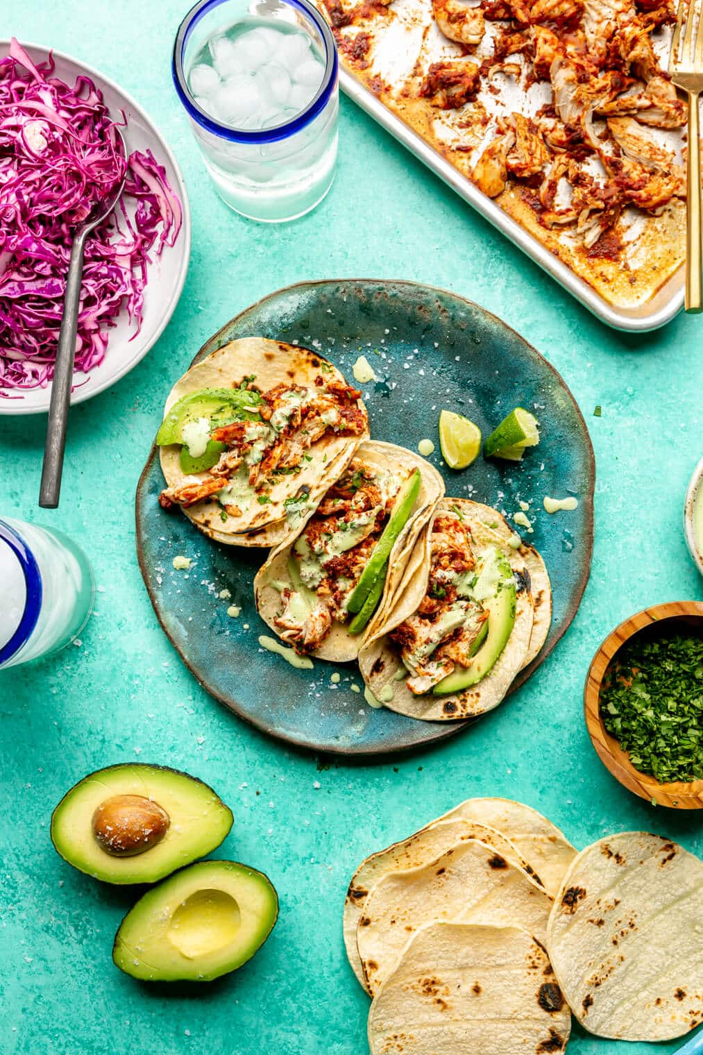 Top down view of three chicken Tinga tacos on a teal plate. The tacos are topped with cilantro, avocado, a drizzle of crema, with a side of lime. There is an avocado cut in half on the table as well as charred corn tortillas, a plate of purple cabbage slaw, a bowl of chopped cilantro, and a sheet pan with chicken Tinga. All are sitting on a teal surface.