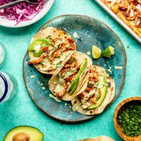 Top down view of three chicken Tinga tacos on a teal plate. The tacos are topped with cilantro, avocado, a drizzle of crema, with a side of lime. There is an avocado cut in half on the table as well as charred corn tortillas, a plate of purple cabbage slaw, a bowl of chopped cilantro, and a sheet pan with chicken Tinga. All are sitting on a teal surface.