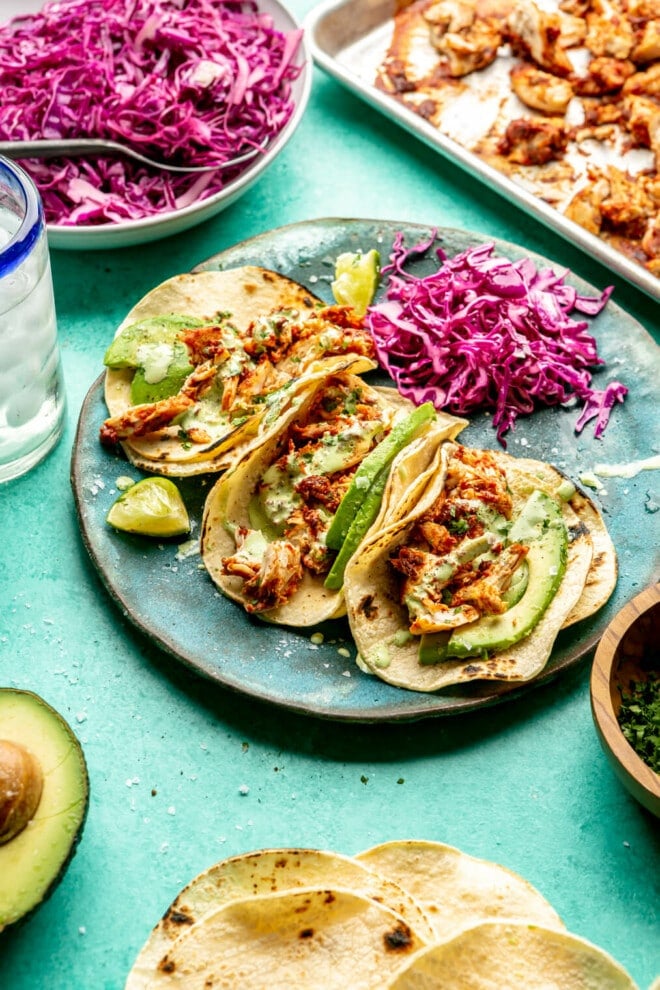 Side view of three chicken Tinga tacos on a teal plate. The tacos are topped with cilantro, avocado, a drizzle of crema, with a side of lime. There is an avocado cut in half on the table as well as charred corn tortillas, a plate of purple cabbage slaw, a bowl of chopped cilantro, and a sheet pan with chicken Tinga. All are sitting on a teal surface.