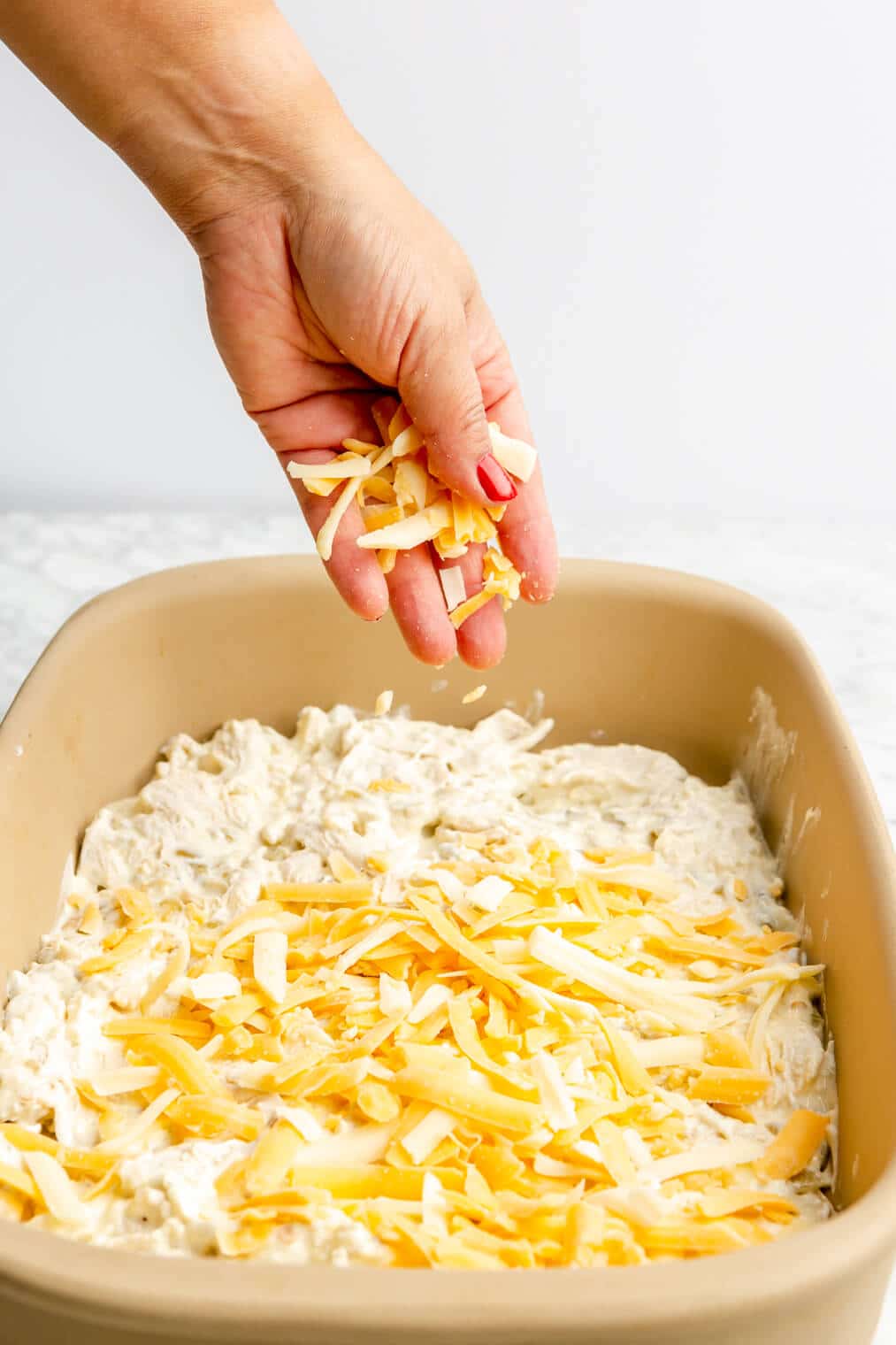 Hand sprinkling shredded cheese on top of the creamy chicken layer in a casserole dish.