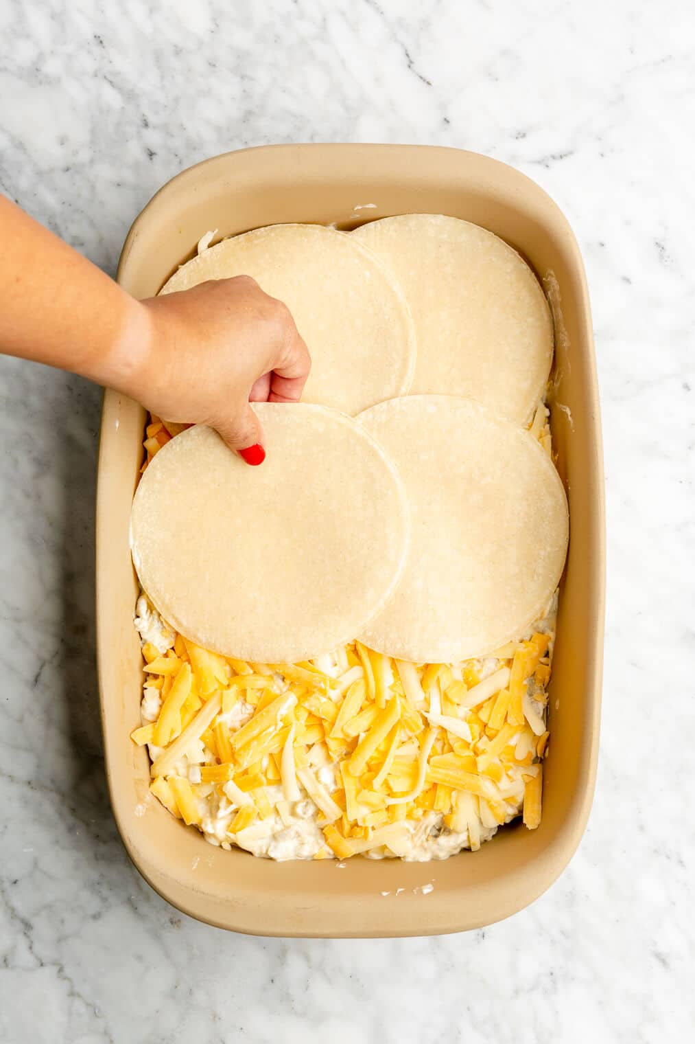 Hand adding another layer of tortillas on top of creamy chicken mixture and shredded cheese in casserole dish.