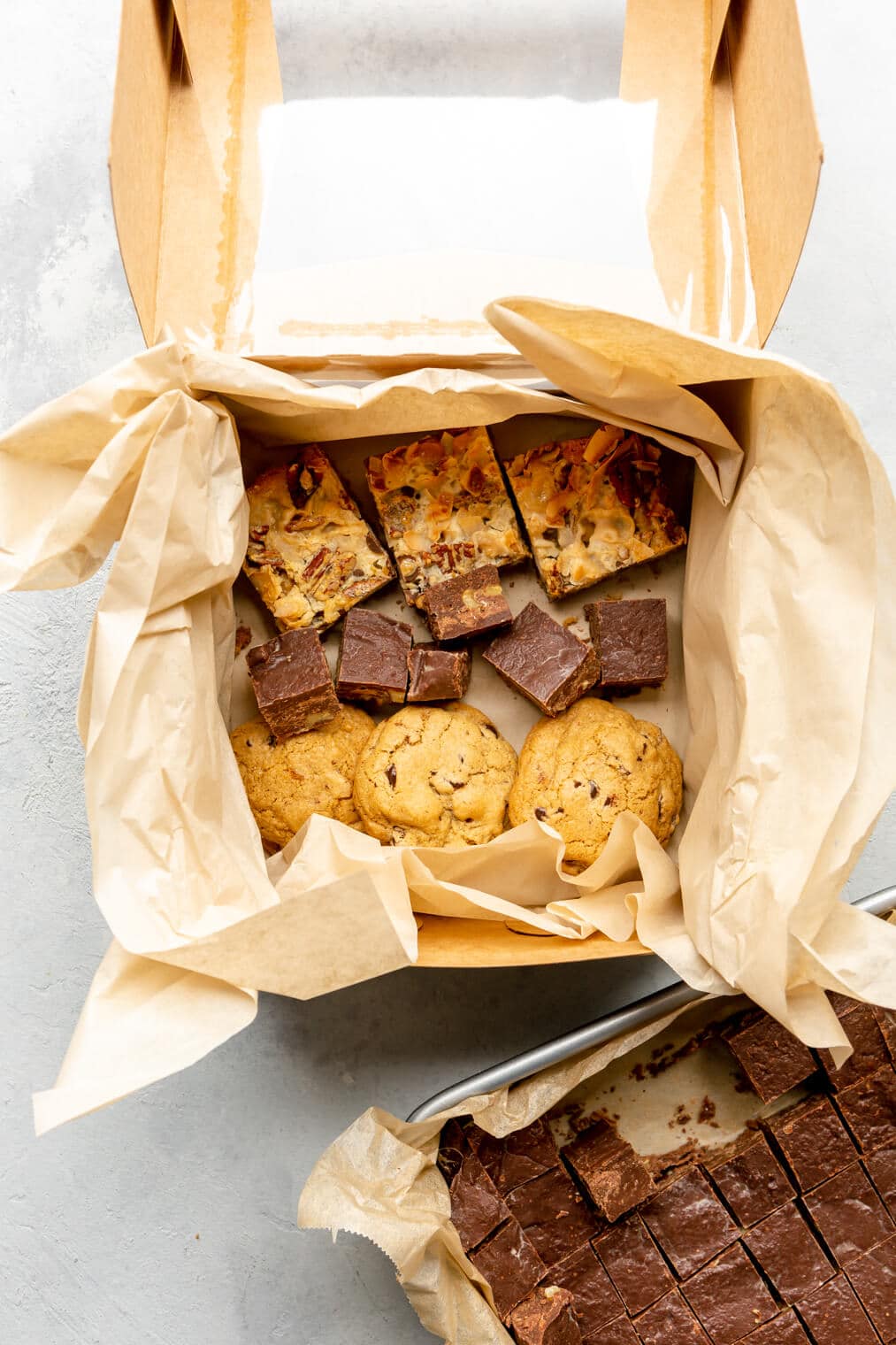 Cookie box lined with parchment paper with rows of cookies, fudge, and bars on the bottom.