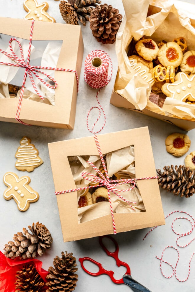 Top down view of three holiday cookie boxes on a grey surface. Two of the cookie boxes are packed and closed and tied with red twine. The third box has the lid open and you can see a variety of cookies in the parchment lined box. There are pine cones, scissors, cookies, and red twine on the surface as well.