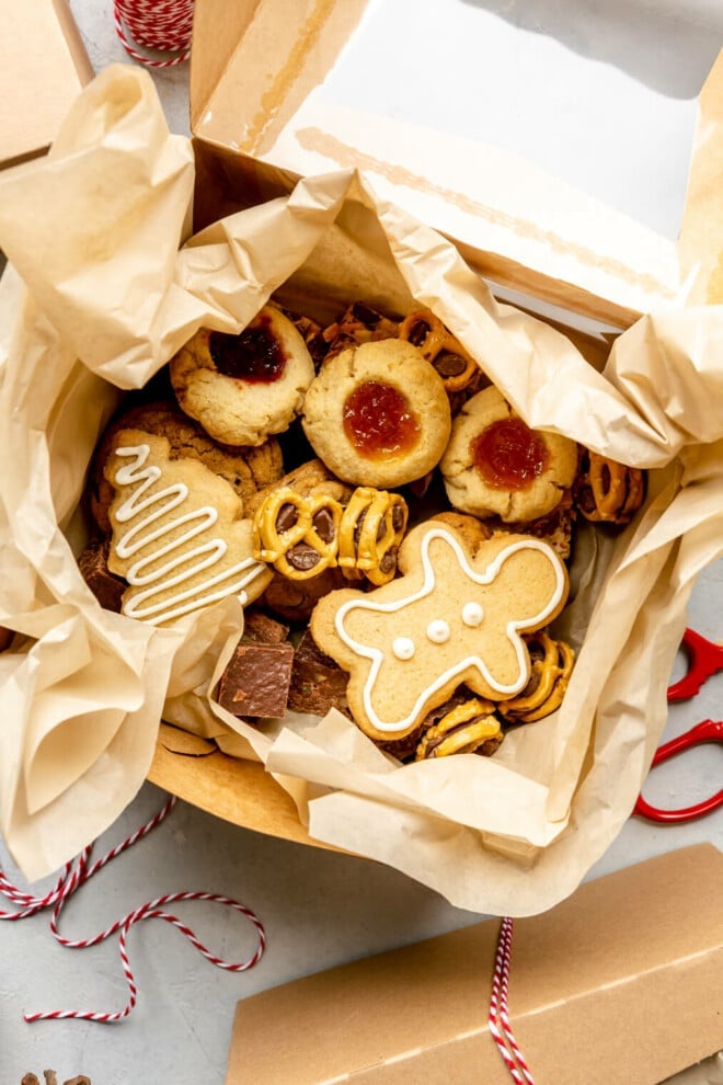 Top down view of a variety of cookies in a parchment paper lined box. The box is on a grey surface and you can see pine cones and red twine on the table.