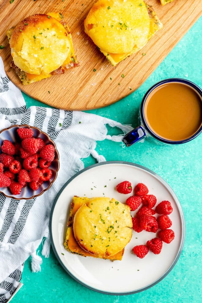 Breakfast sandwich on a plate with a side of raspberries. There is a bowl of raspberries and blue rimmed mug with coffee and a wooden cutting board with 2 breakfast sandwiches also on the table. 