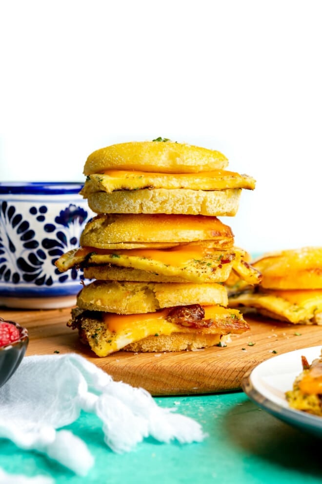 Stack of 3 breakfast sandwiches on a wooden cutting board. There is a white and blue mug in the background.