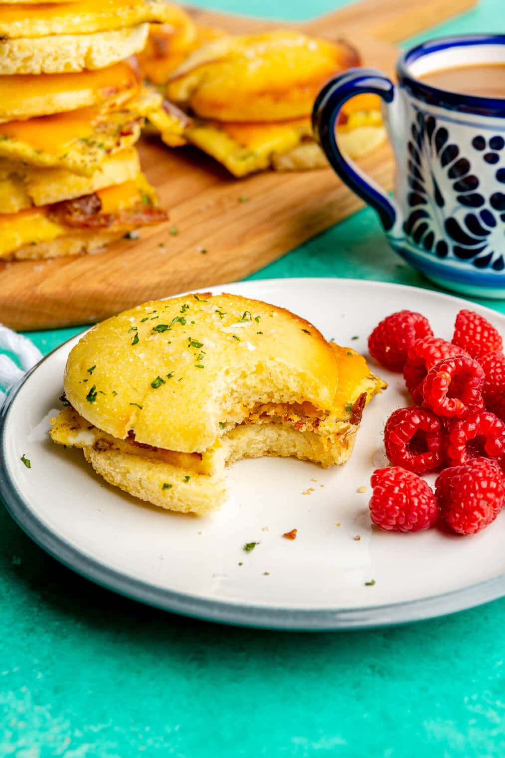 Breakfast sandwich on a plate with a side of raspberries. There is a white and blue mug in the background and a wooden cutting board with other breakfast sandwiches on the board. All are on a teal surface.