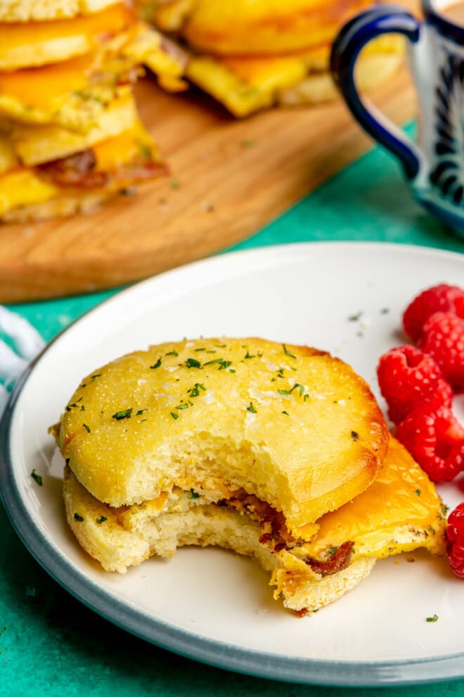Breakfast sandwich on a plate with a side of raspberries. There is a white and blue mug in the background and a wooden cutting board with other breakfast sandwiches on the board. All are on a teal surface.