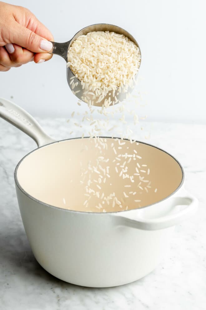 Hand pouring rice into a white pot from a measuring cup.