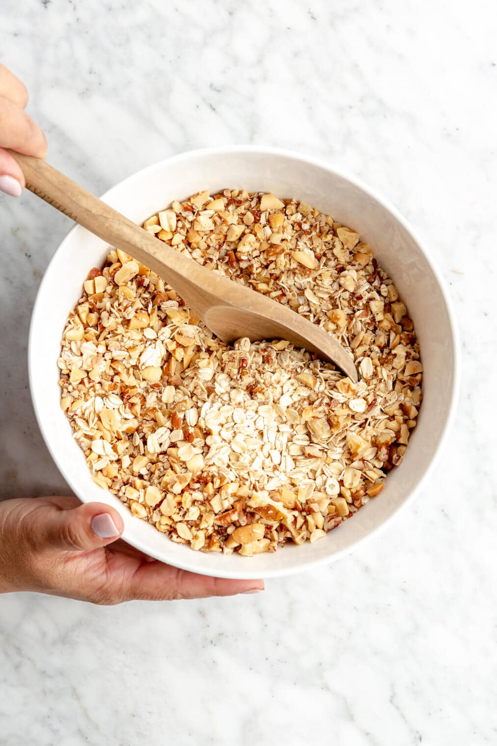Hand holding the side of a white bowl and the other hand is holding a wooden spoon and stirring dry oats and nuts together.