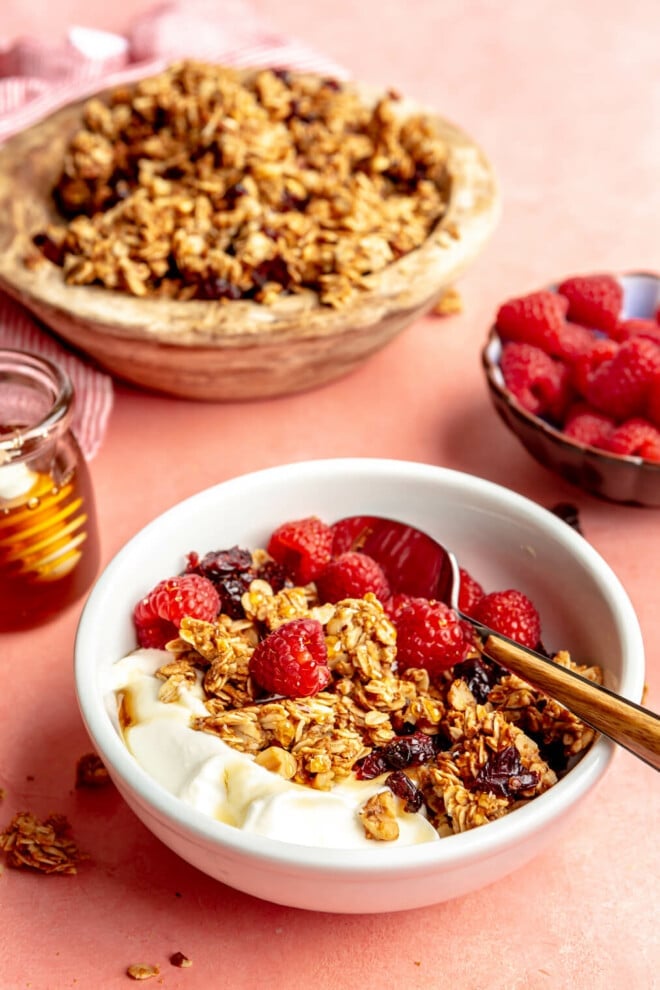 White bowl with yogurt and granola topped with raspberries. There is a wooden bowl of granola in the background and a bowl of raspberries. All are sitting on a pink blush surface. 