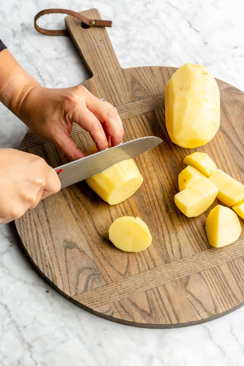Hand holding a knife and cubing potatoes on a wooden cutting board.