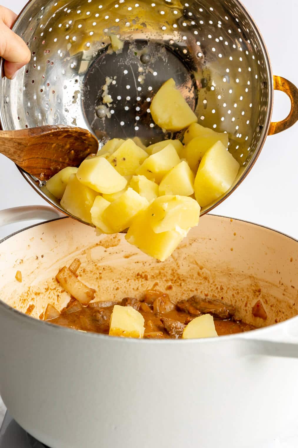 Wooden spoon adding boiled potatoes to a white dutch oven.