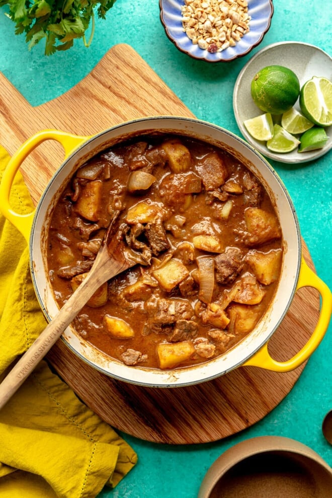 Top down view of beef massaman in a yellow handled dutch oven and a wooden spoon resting inside. The dish is sitting on top of a wooden cutting board and there is a plate of sliced limes in the top corner. All are sitting on a bright teal surface. 