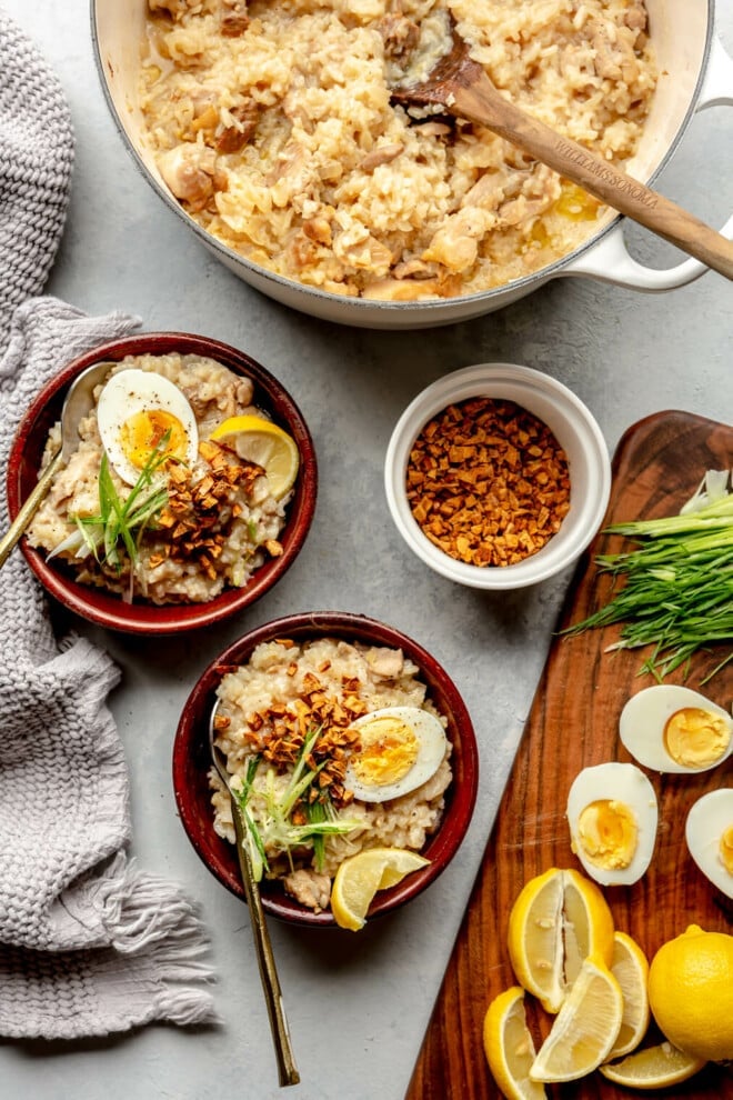 Top down view of a grey tablescape set with two wooden bowls of arroz clad and a large pot with arroz caldo. There is a white dish with crispy fried garlic and a wooden cutting board with hard-boiled eggs, lemon wedges, and sliced green onions. 