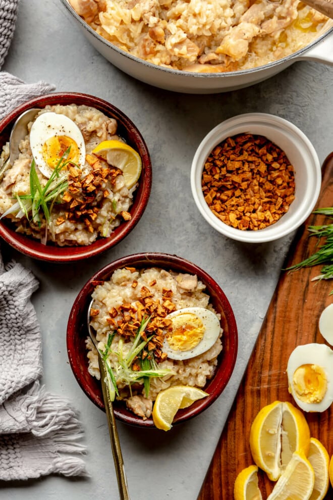 Top down view of a grey tablescape set with two wooden bowls of arroz clad and a large pot with arroz caldo. There is a white dish with crispy fried garlic and a wooden cutting board with hard-boiled eggs, lemon wedges, and sliced green onions.