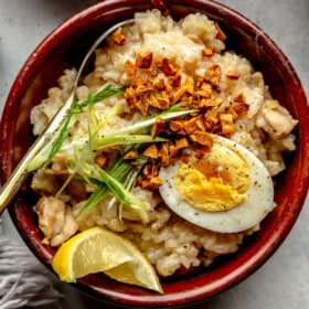 Top down view of a wooden bowl filled with chicken arroz clad. The rice congee is topped with a hard boiled egg, crispy garlic, sliced green onion, and a lemon wedge.