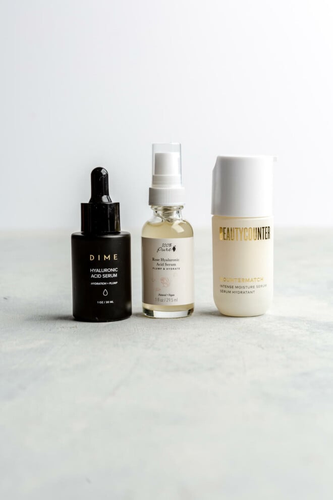 Three hyaluronic acid serums lined up on a marble surface with a white background. One of the bottles is black with a dropper. One is clear with a white label and a pump, and the other is in a frosted white container with a white lid with a pump.