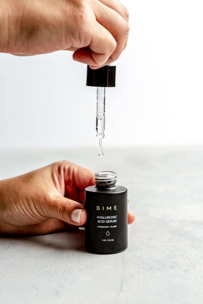 Hand holding a Dime serum container in one hand and a dropper in another. There is a drop of serum falling from the dropper. 