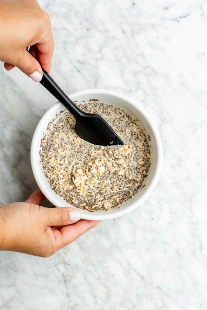 Hand holding a small, black spatula stirring oats, chia, and milk in a bowl.