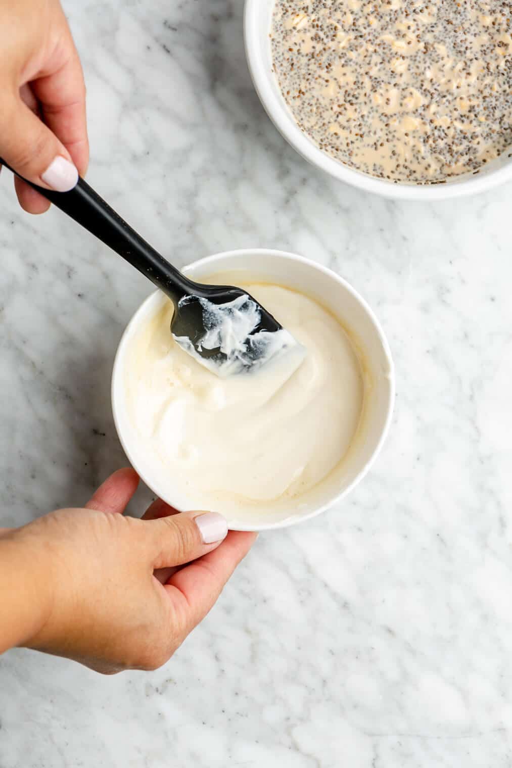 Hand using a black spatula to stir yogurt and maple syrup together in a small white bowl.