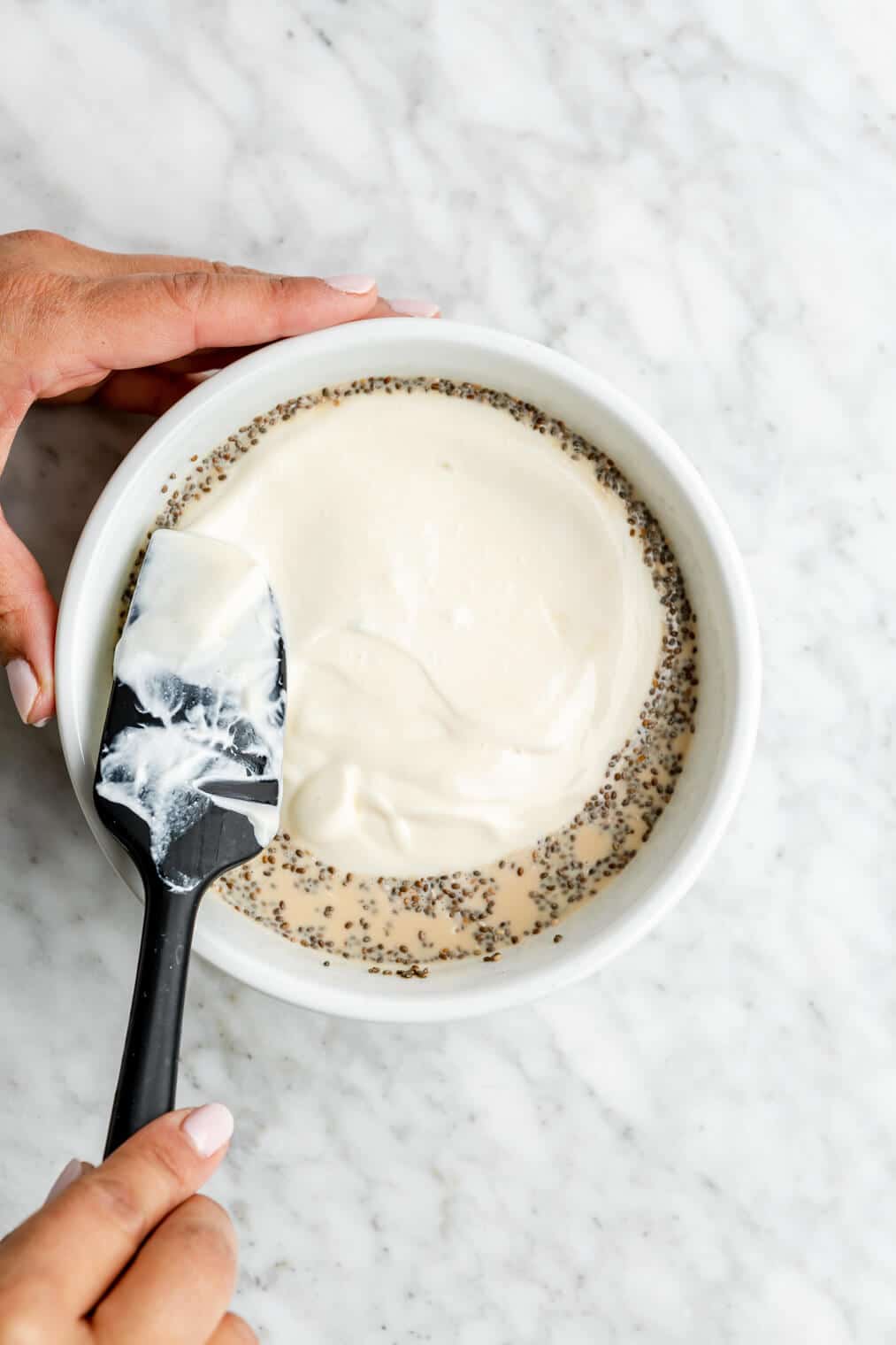 Hand using a black spatula to spread yogurt over the top of the oat mixture in a bowl.