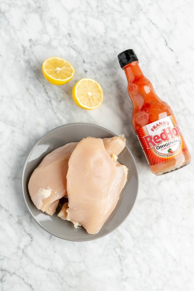 Buffalo chicken marinade ingredients on a grey and white marble surface. There's a bottle of Frank's red hot sauce and a lemon sliced in half and a plate with two chicken breast. 
