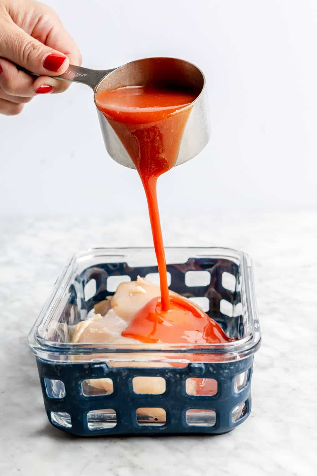 Hand holding a stainless steel measuring cup pouring hot sauce over a chicken breast in a glass container lined with blue netted silicone.