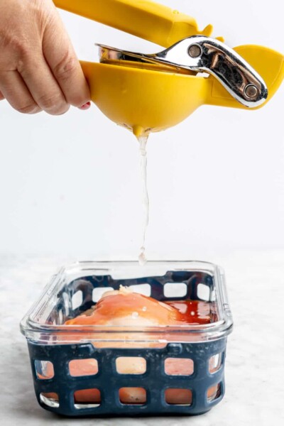 Hand squeezing a citrus juicer over top a chicken breast coated in hot sauce in a glass container with a blue netted silicone liner on the outside.