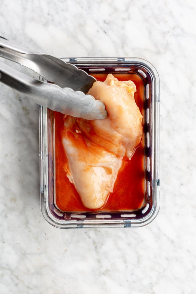Pair of tongs lifting a chicken breast out of buffalo marinade in a glass container.