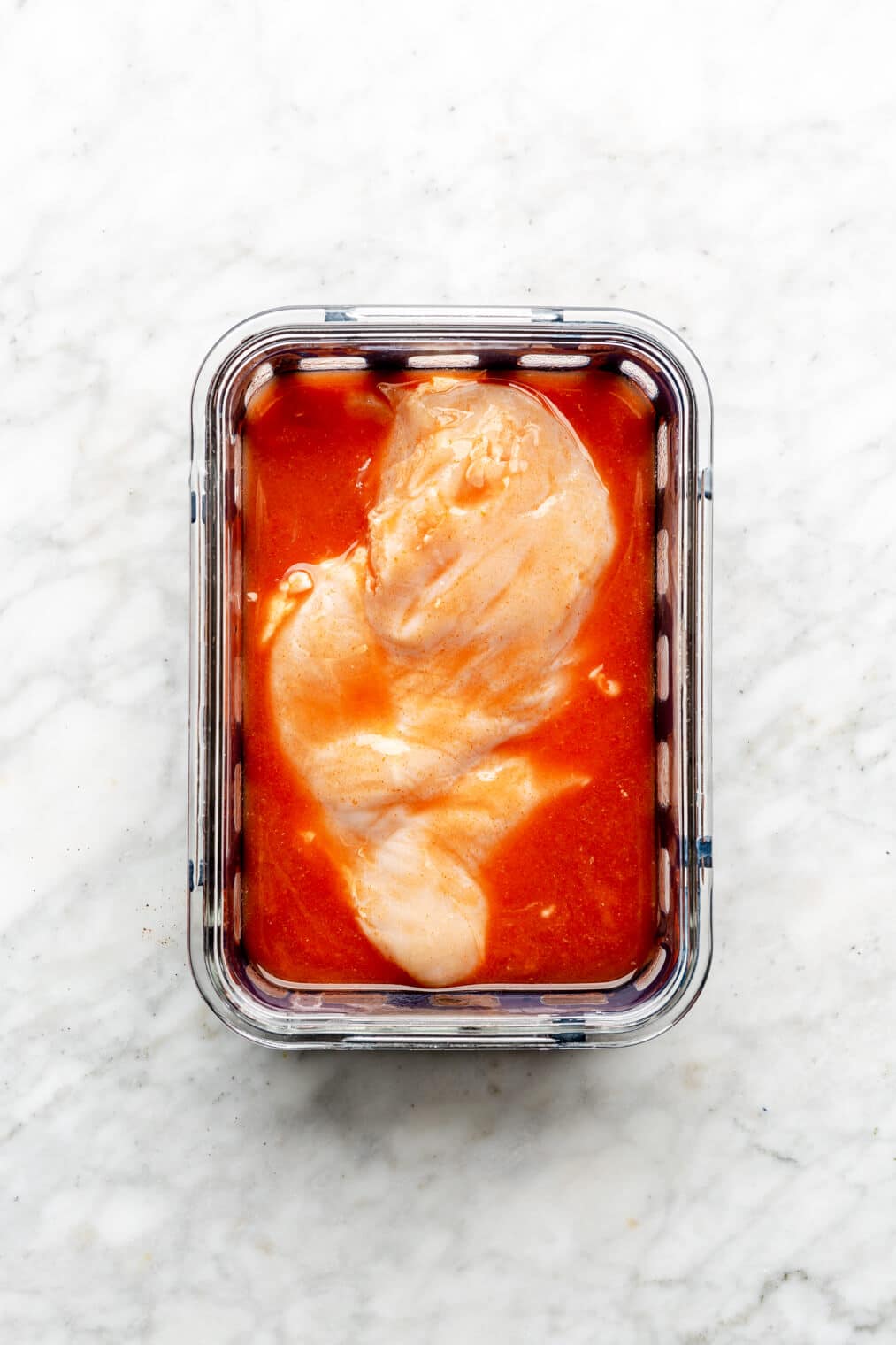 Chicken breast marinating in buffalo marinade in a glass container.