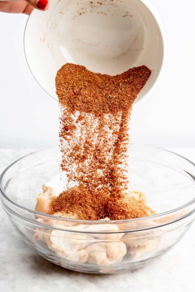Dry rub being poured over a bowl of chicken wings in a large glass bowl.