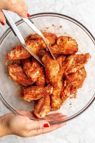 Hand holding a pair of tongs and tossing chicken wings in a dry rub spice mixture.
