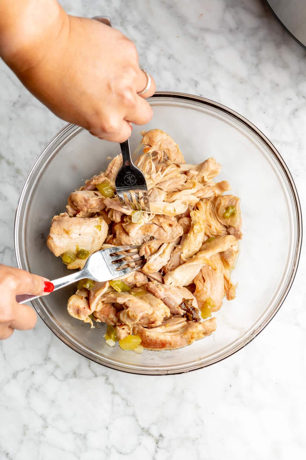 Hands using two forks to shred chicken in a glass bowl.