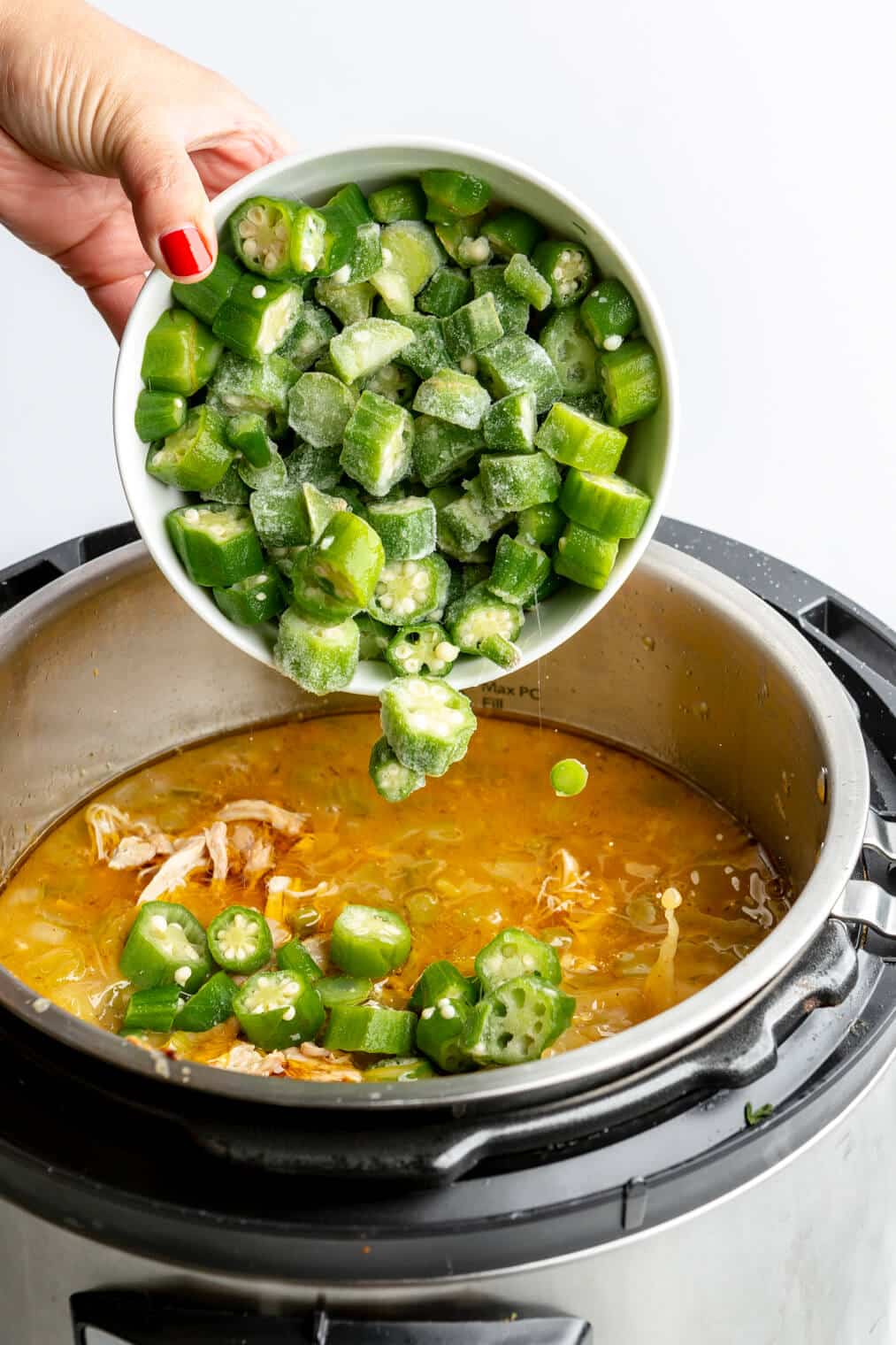 Hand holding a white bowl with frozen okra in it and pouring it into the instant pot.