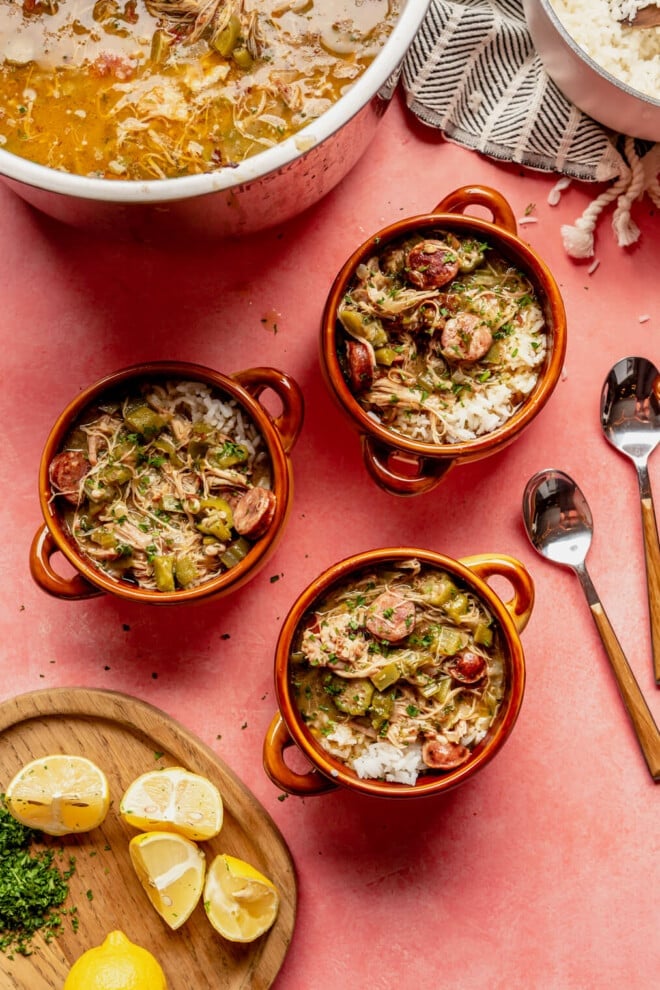 Top down view of three bowls of gumbo in little orange serving pots with small handles. They are on a pink colored table with a cutting board with lemons and fresh herbs. There is also a pot of gumbo in the background.