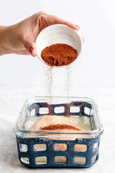 Hand holding a small white bowl pouring chili powder over a chicken breast with lime juice in a glass container with blue netted silicone.