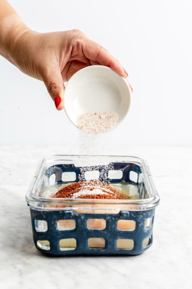 Hand pouring salt over top of a chicken breast with chili powder and lime juice in a glass container with blue netted silicone.