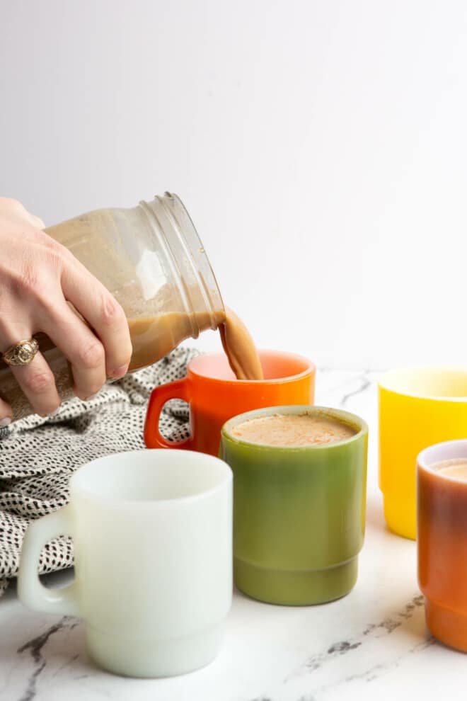 Hand holding mason jar of mushroom coffee, pouring a cup of coffee on a table with 4 other mugs. 