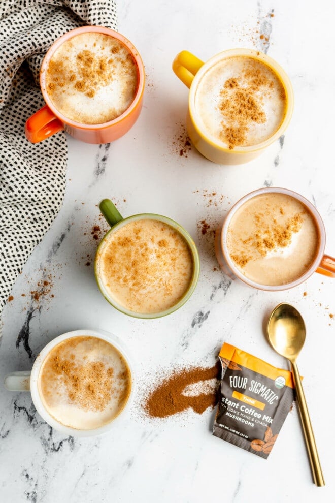 5 cups of mushroom coffee on a black and white marble surface. There is a grey linen to the left of the mugs and a packet of Four Sigmatic mushroom coffee on the bottom right corner next to a gold spoon. 