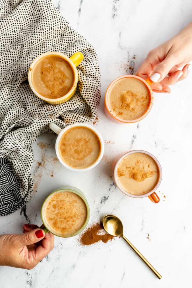 5 cups of mushroom coffee on a black and white marble surface. Two hands are reaching in and holding the handle of two of the cups. There is a grey linen and gold spoon with a sprinkle of mushroom coffee underneath the spoon.