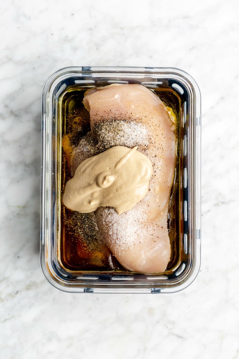 Chicken breast with honey mustard marinade ingredients on top of the chicken breast in a glass container on a grey and white marble surface.