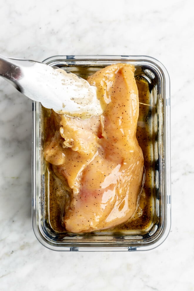 Tongs holding a chicken breast being tossed in honey mustard marinade in a glass container on a grey and white marble surface.