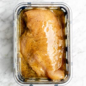 Honey mustard marinated chicken breast in a glass container on a grey and white marble surface.