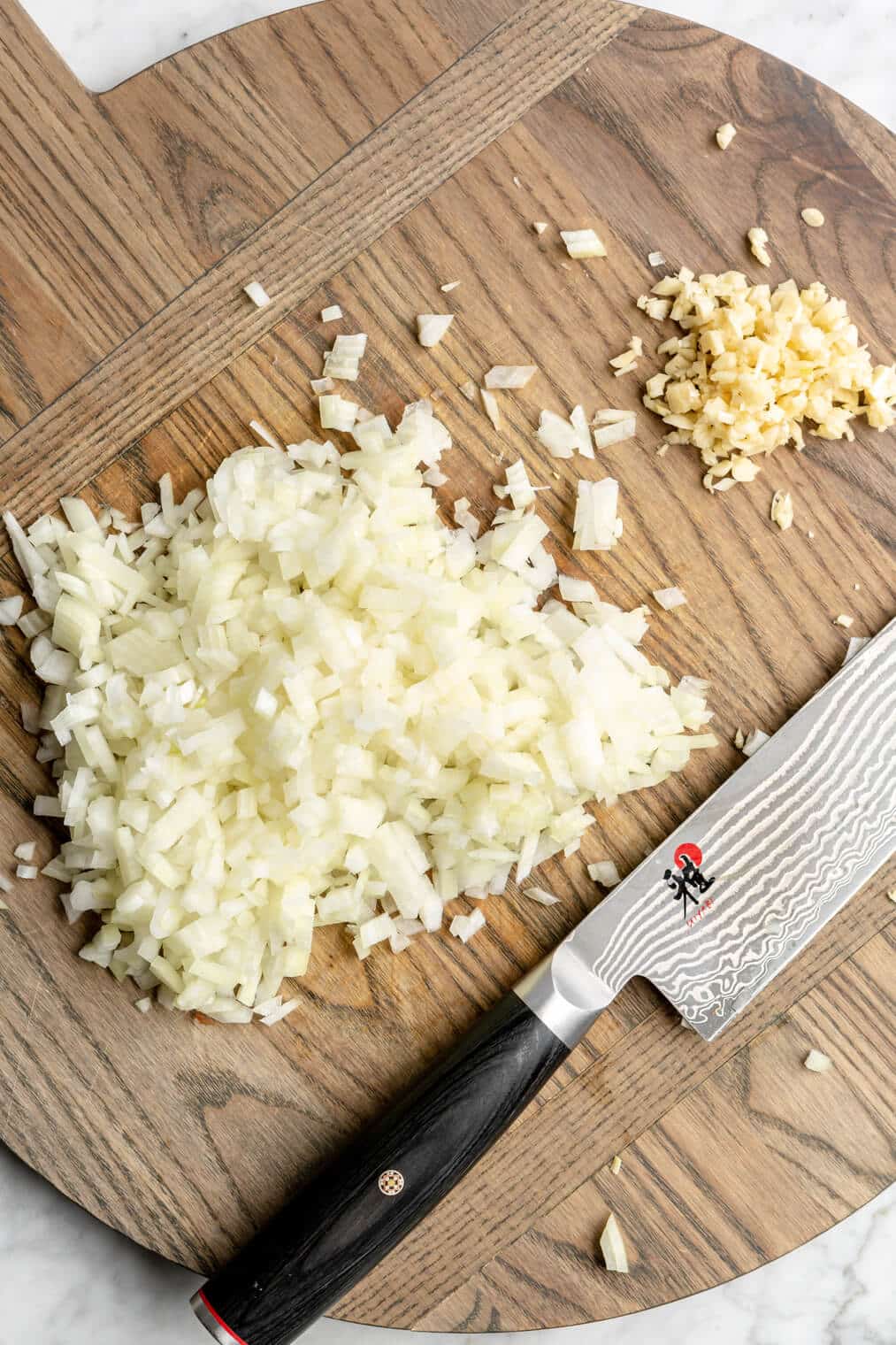 There is a wooden cutting board with diced onion and minced garlic with a chef's knife.