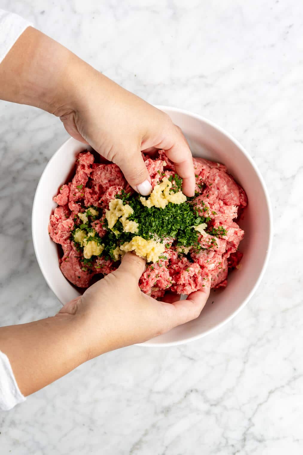 Hands mixing together ground beef with garlic and parsley in a white bowl.