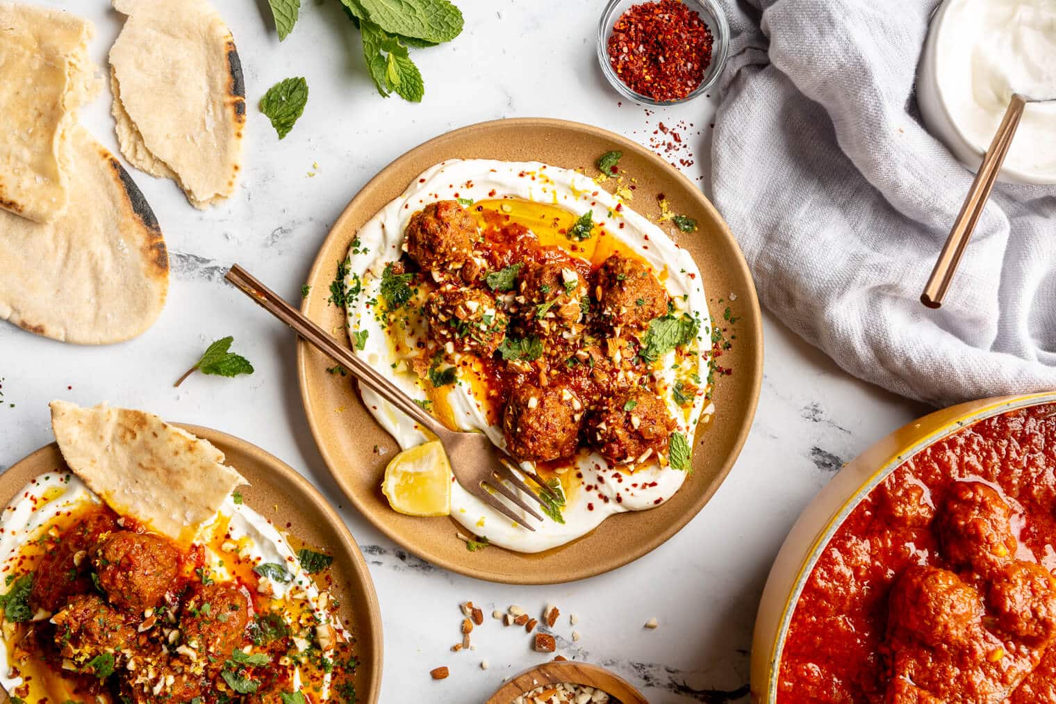 A plate of Moroccan-inspired meatballs on a bed of greek yogurt and garnished with parsley, lemon, and sauce on a white and black marble surface. There are basil leaves, pita breads, a linen, and another plate of meatballs on the table.