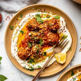 Top down view of a brown plate with a serving of Moroccan-inspired meatballs sitting on a spread of white, Greek yogurt garnished with Aleppo flakes, sauce, parsley, and lemon.
