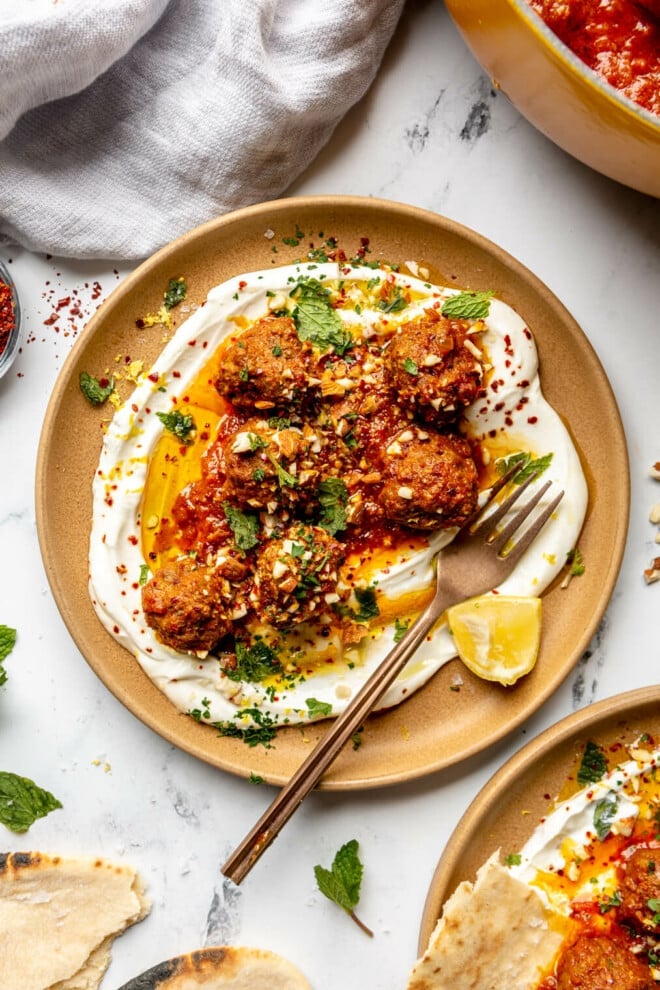 Top down view of a brown plate with a serving of Moroccan-inspired meatballs sitting on a spread of white, Greek yogurt garnished with Aleppo flakes, sauce, parsley, and lemon. 